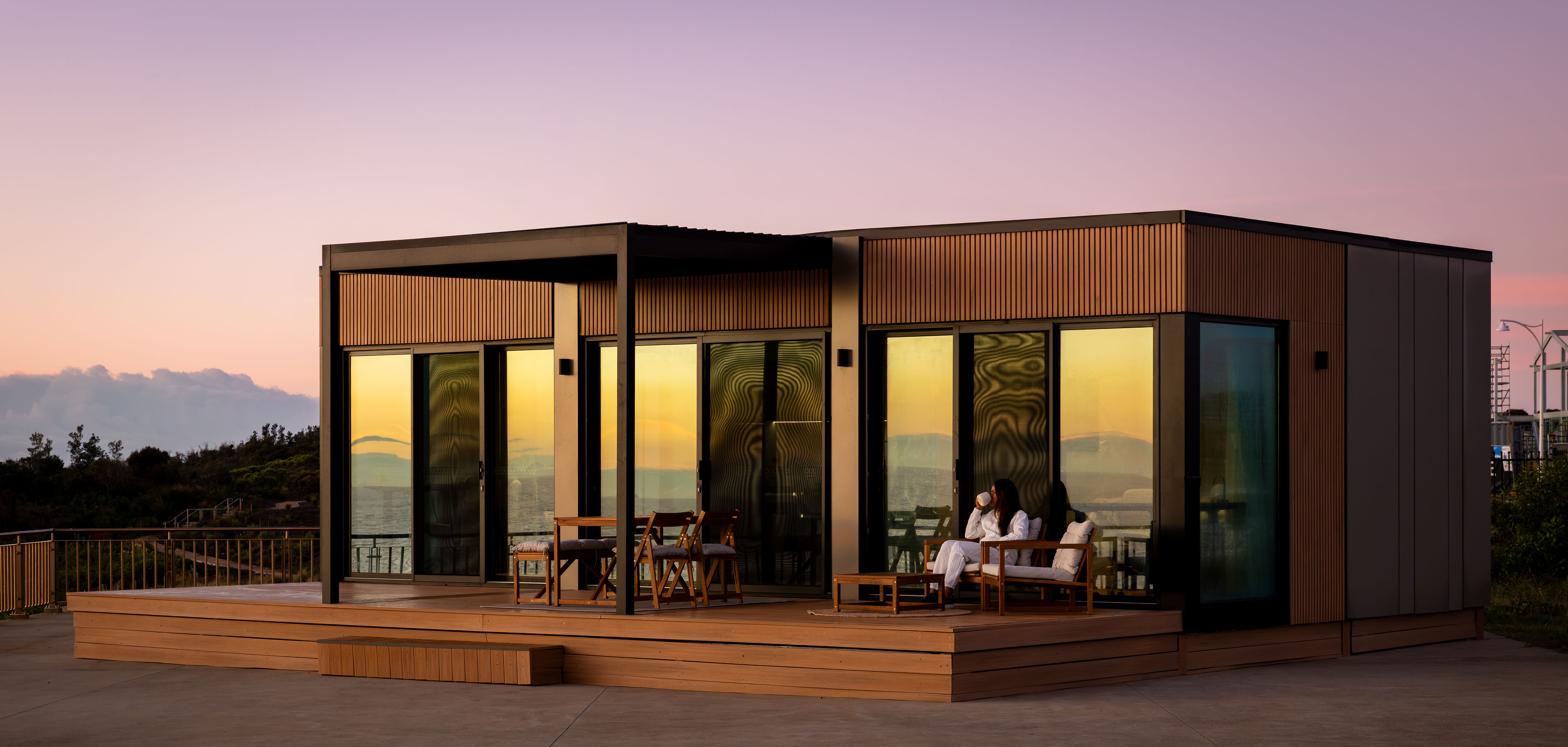OHOME at Catherine Hill Bay at dusk, glass glowing, a woman on the deck with a coffee